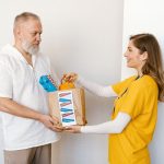Homem idoso recebe uma cesta de doações de uma voluntária sorridente em um ambiente interno.