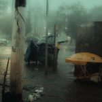 Rua urbana à noite sob forte chuva, com carros em circulação e pessoas protegidas por guarda-chuvas.