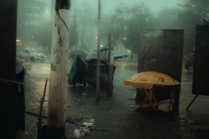 Rua urbana à noite sob forte chuva, com carros em circulação e pessoas protegidas por guarda-chuvas.
