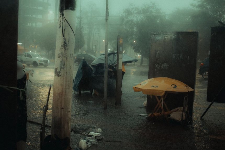 Rua urbana à noite sob forte chuva, com carros em circulação e pessoas protegidas por guarda-chuvas.