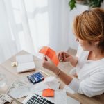 Mulher organiza contas e recibos em uma mesa, usando calculadora e laptop para planejar as finanças pessoais.