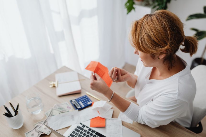 Mulher organiza contas e recibos em uma mesa, usando calculadora e laptop para planejar as finanças pessoais.
